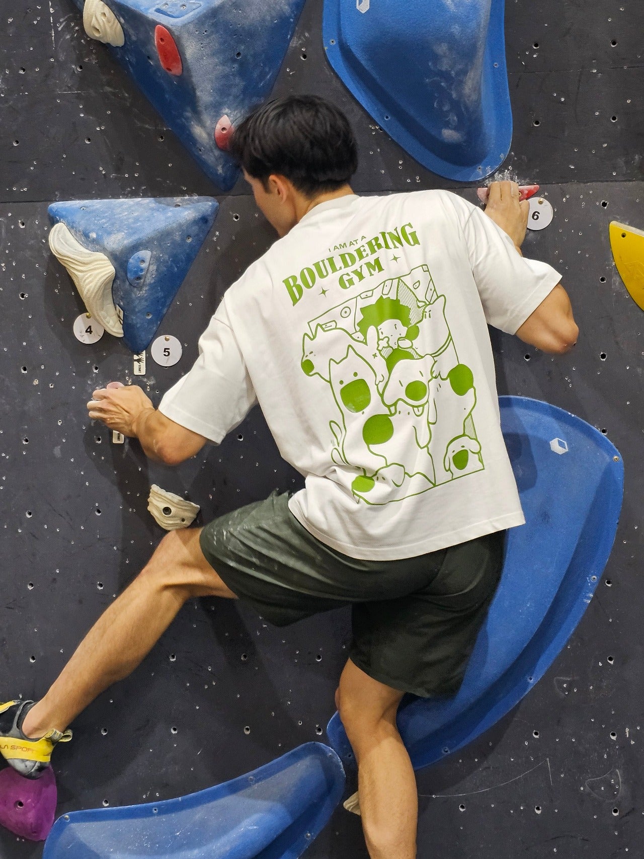 man wearing thick white nobori tee shirt climbing in indoor bouldering gym with back faced to camera
