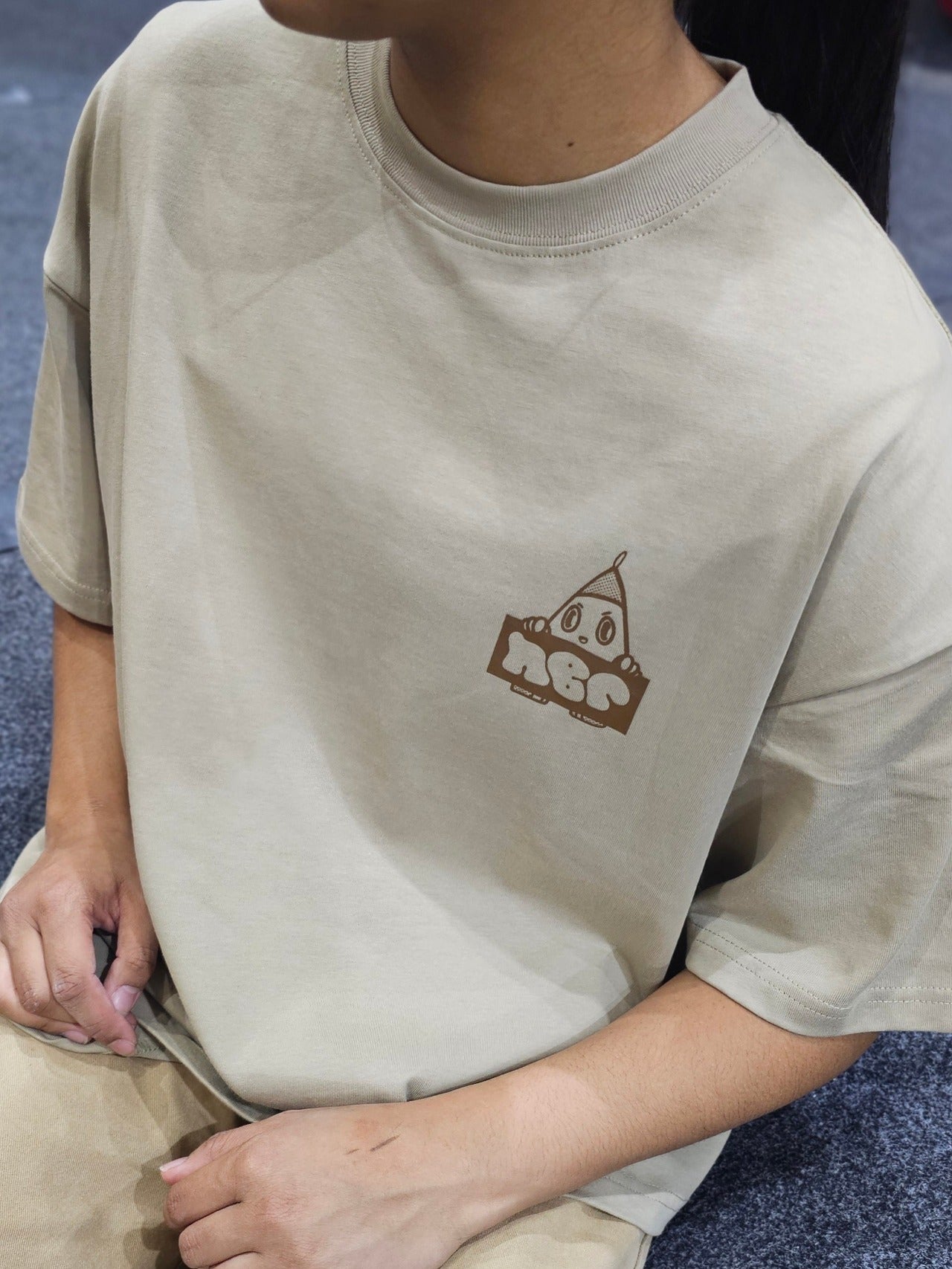 Girl sitting down on mat inside bouldering gym wearing brown oversized nobori tee shirt facing the camera